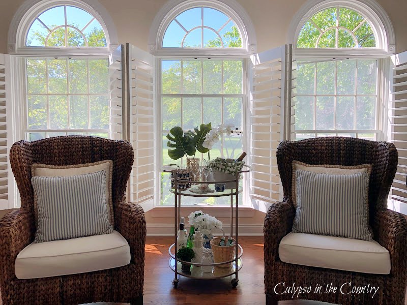 seagrass chairs and tropical bar cart in front of three open windows with plantation shutters