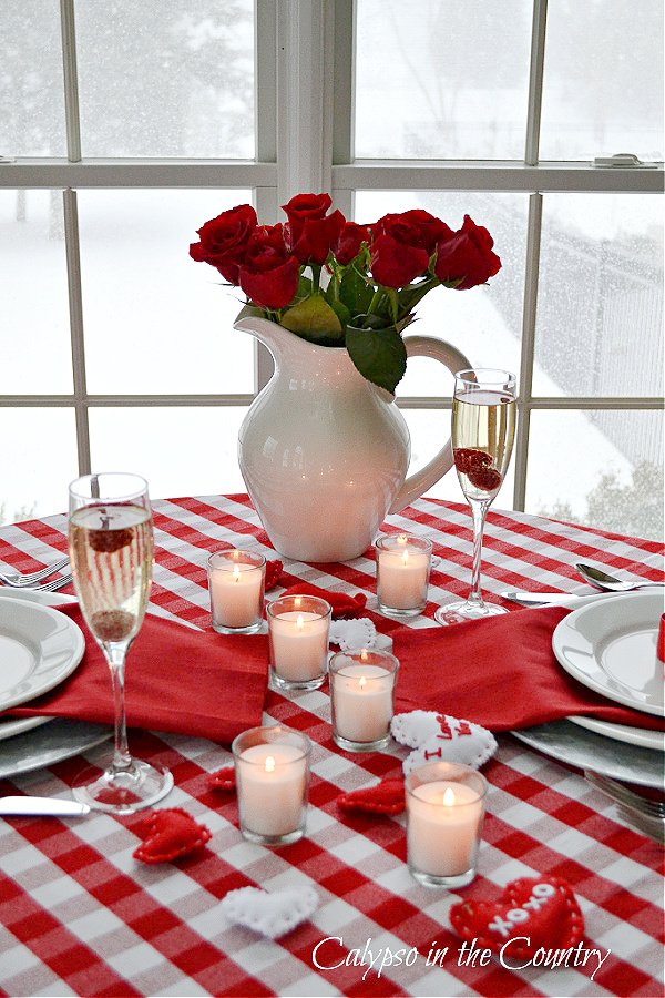 Red roses in white pitcher on red and white checked table cloth with white candles - valentines day tablescape