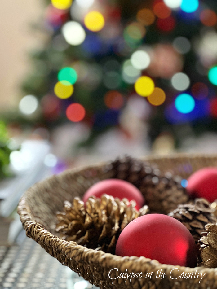 Red ball ornaments and pine cones in round basket with Christmas lights in background - goodbye December hello January