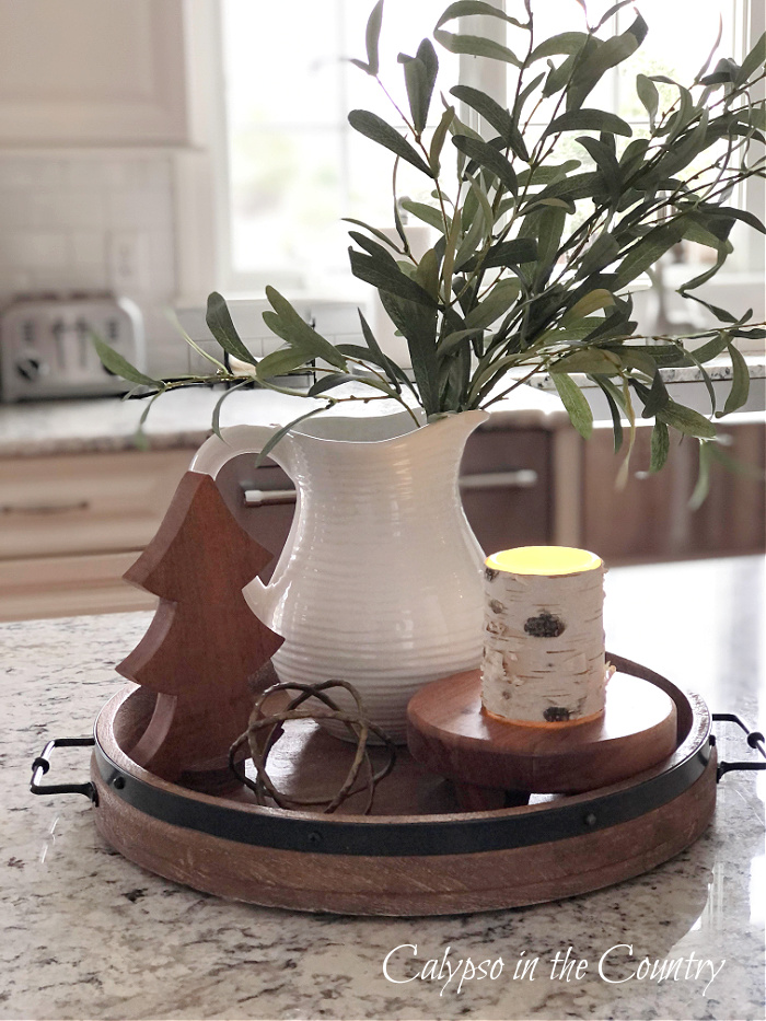 Round wooden tray with white pitcher of greenery, a wooden tree and birch candle on stand. - January decorating inspiration 