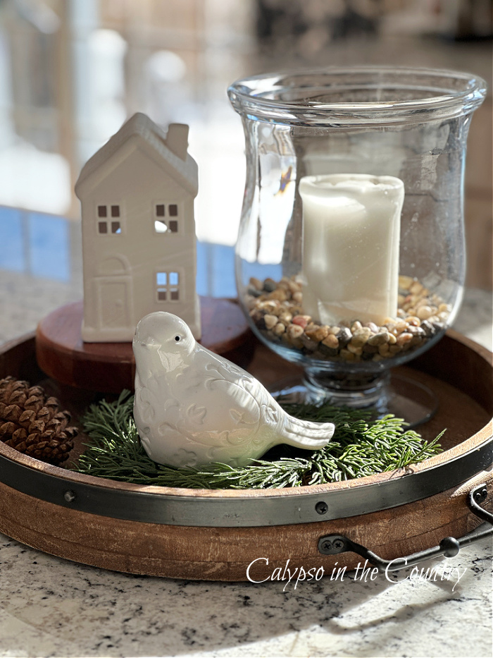 White ceramic house and bird with white candle in glass hurricane on round wooden tray 