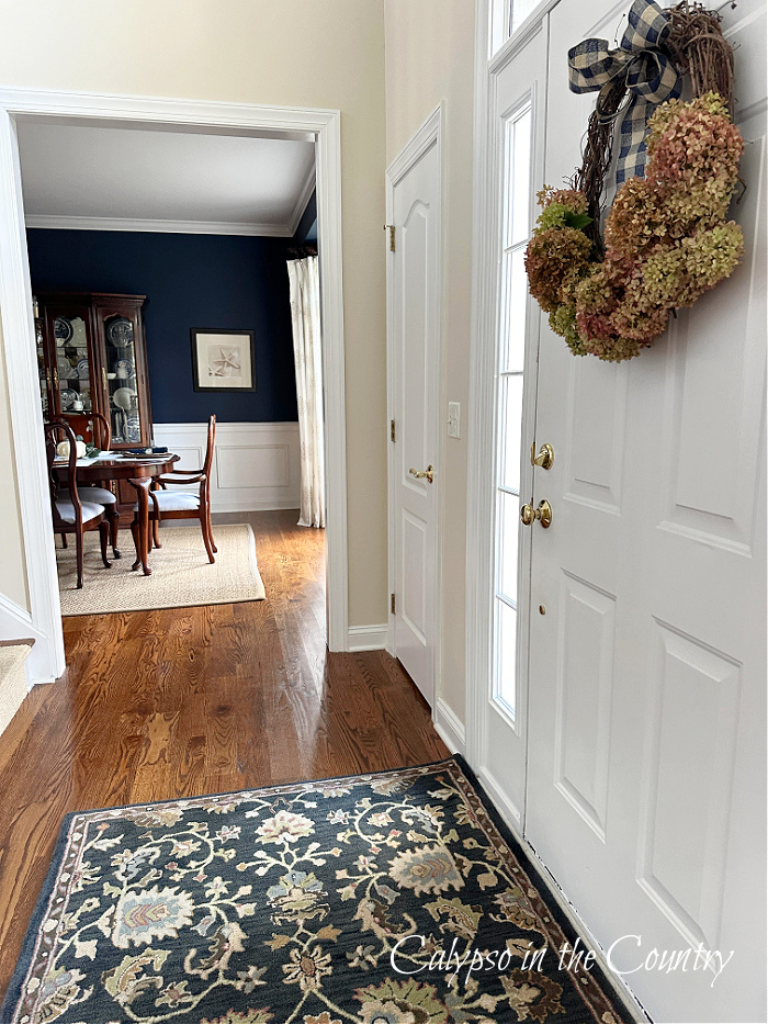 Foyer with white door and blue rug with hydrangea wreath hanging on door - fall feeling