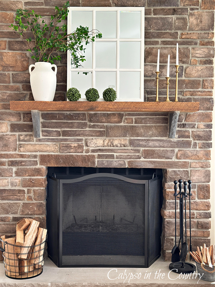 stone fireplace with window pane mirror, white vase filled with faux green stems, gold candlesticks and three faux boxwood spheres