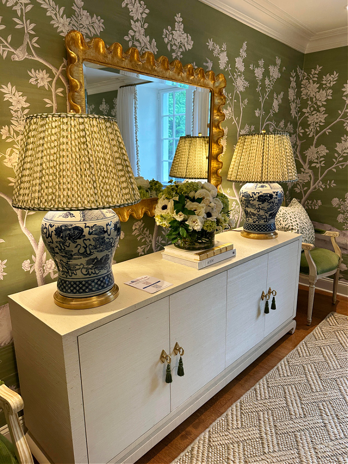Two blue and white porcelain lamps with pleated shades on white buffet table with gold mirror above and green floral wallpaper - may recap