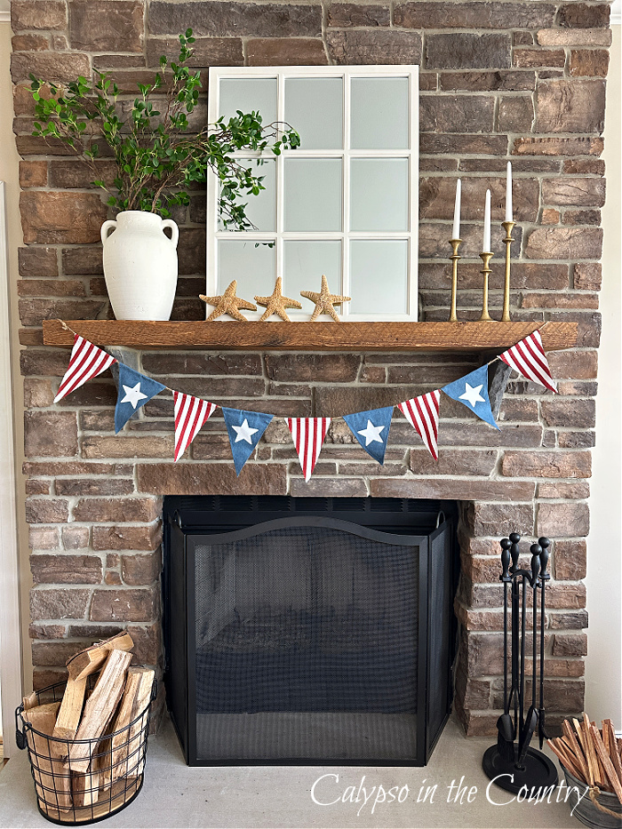 stone fireplace decorated with window pane mirror, white vase of greenery and a red white and blue hanging banner