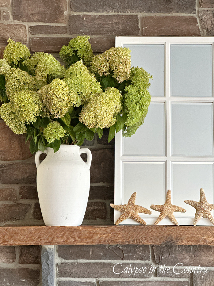 White vase filled with green hydrangeas next to white windowpane mirror on wood mantel on stone fireplace - decorating with hydrangeas from summer to fall