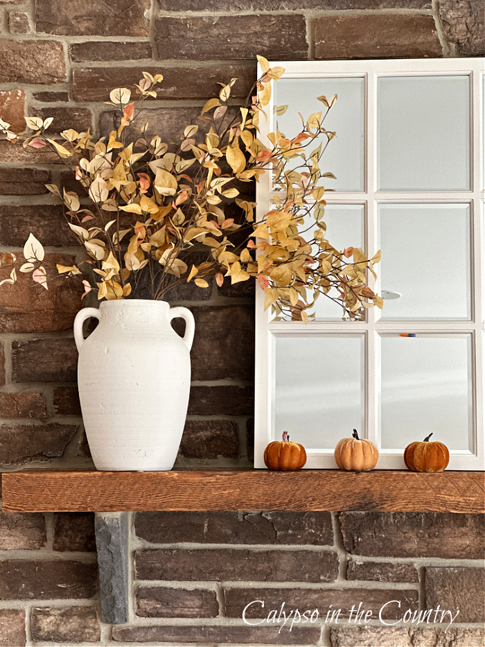 Vase of golden fall leaves next to white window pane mirror on mantel of stone fireplace