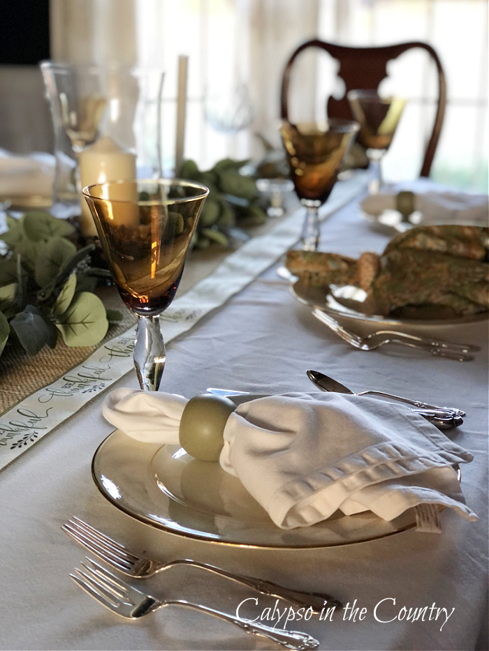 Place settings on table with white table cloth, burlap table runner, eucalyptus and printed ribbon - Thanksgiving table runner ideas