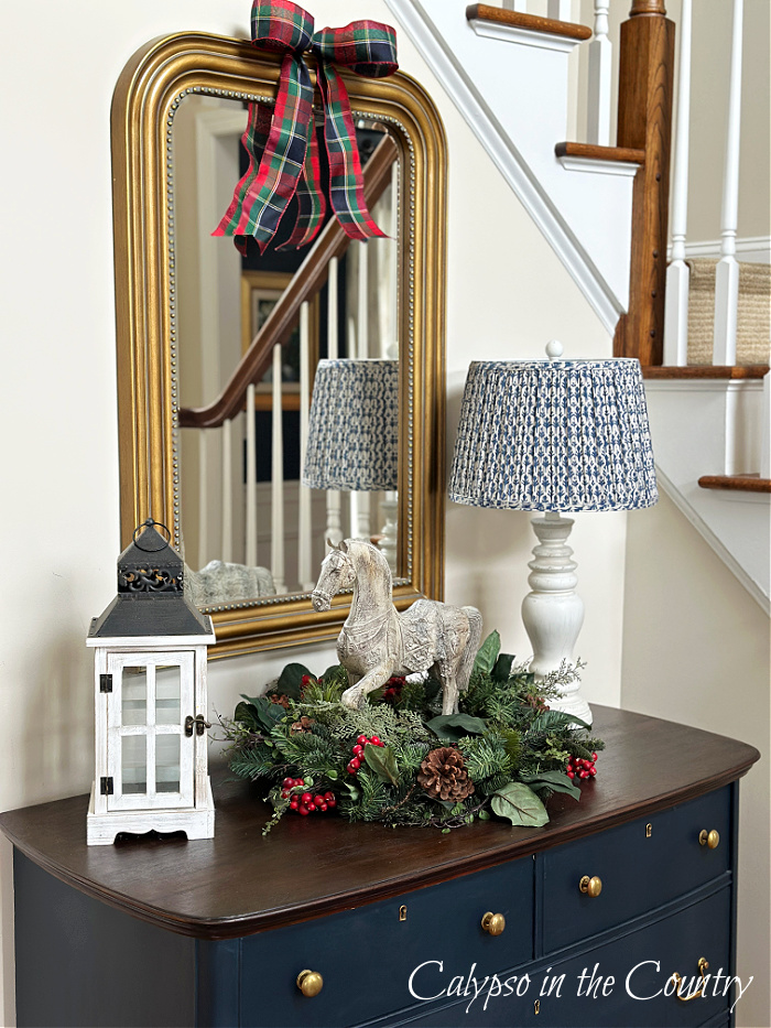 Blue dresser with lamp, horse statue and wreath under gold mirror by staircase