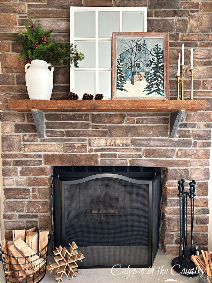 Stone fireplace decorated for winter with white vase filled with greenery, white mirror and snowy painting 