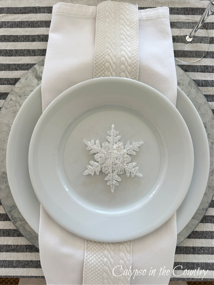 Layered white plates and white napkin with sparkly snowflake ornament on top
