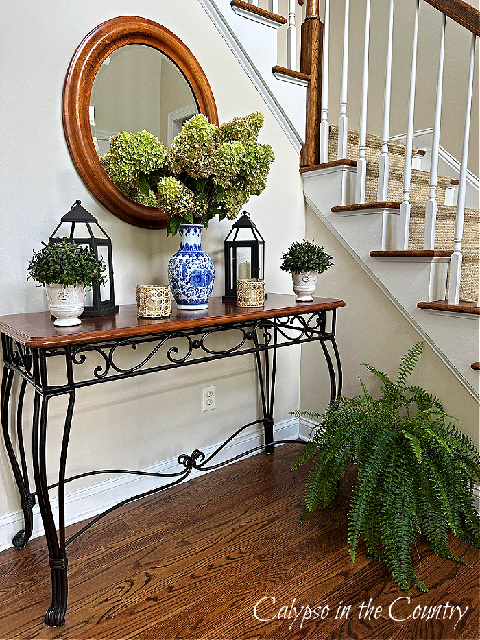 Foyer table with blue and white vase of hydrangeas with a boston fern plant on floor next to stairs - fern decorating ideas 