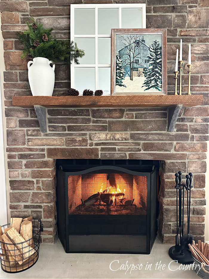 Wood burning fire in stone fireplace with mantel decorated with greenery, white paned mirror and winter artwork