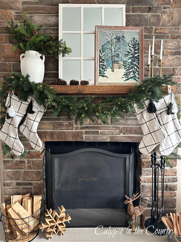 Stone fireplace with greenery in white vase next to white mirror and winter artwork above draped garland and white Christmas stockings- Decorating a mantel for every season