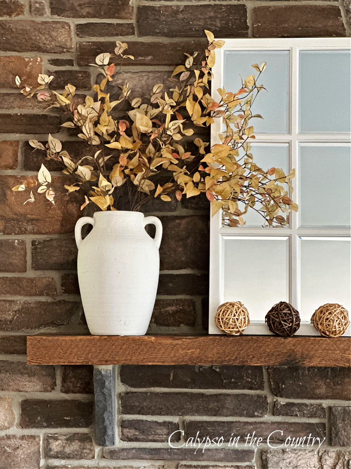 Stone fireplace with golden leaves in a white vase, a white paned mirror and wicker orbs - How to decorate a mantel for every season
