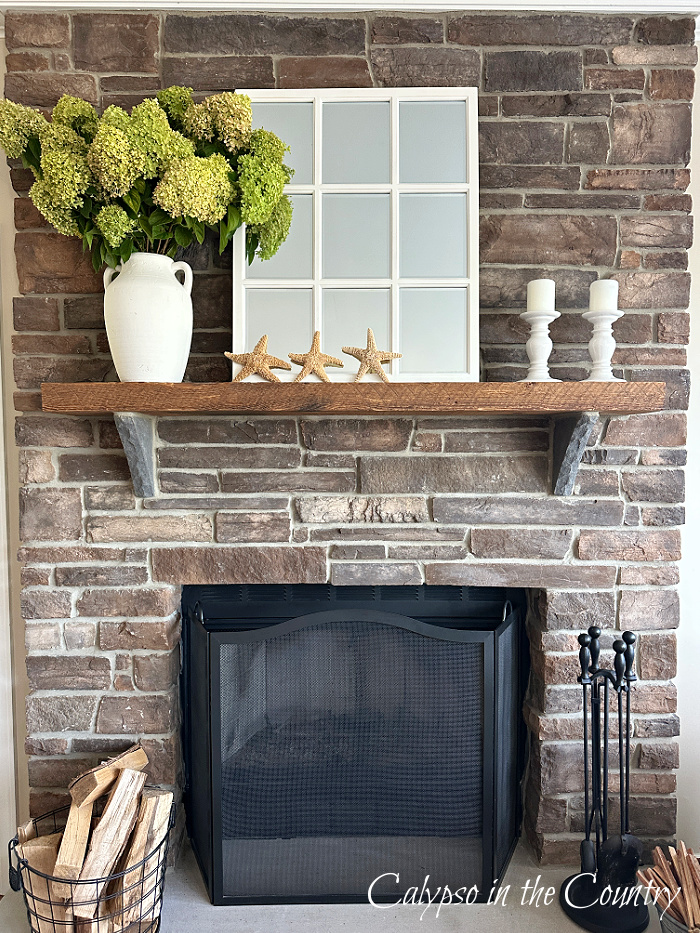Stone fireplace with green hydrangeas in a white vase, a white paned mirror, starfish and white candles - How to decorate a mantel for every season