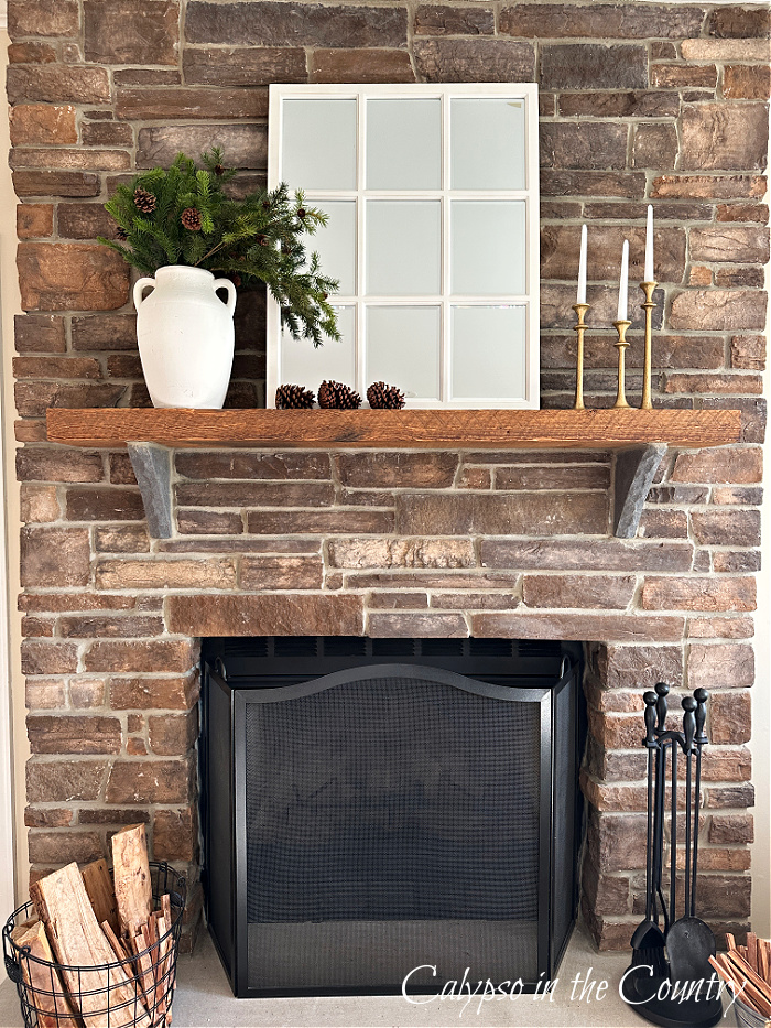 Stone fireplace with white vase of greenery, white paned mirror, candles and pine cones - decorate a mantel for every season