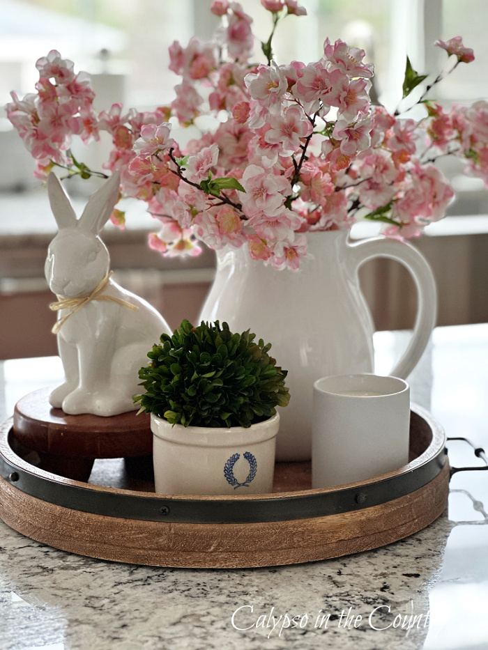 White ceramic bunny and white pitcher of pink cherry blossoms on round wooden tray