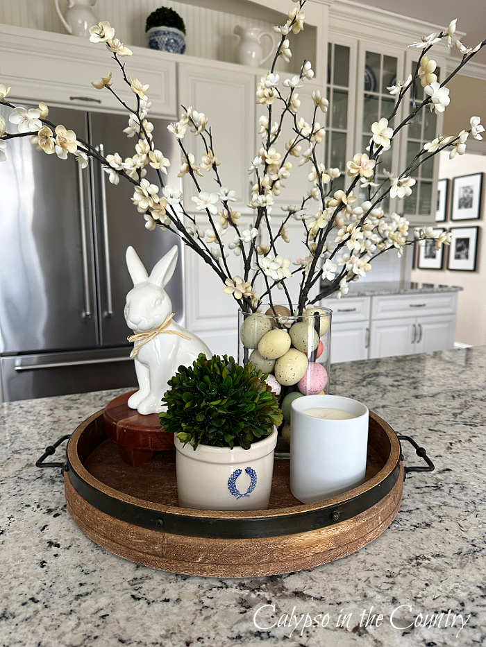 Round tray on counter holding white bunny, greenery and vase with Easter eggs and white dogwood branches