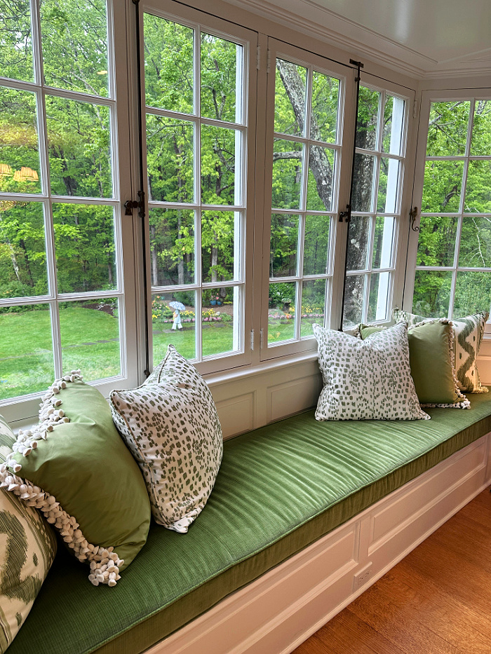 Large paned windows overlooking yard with a green velvet window seat with green and white pillows