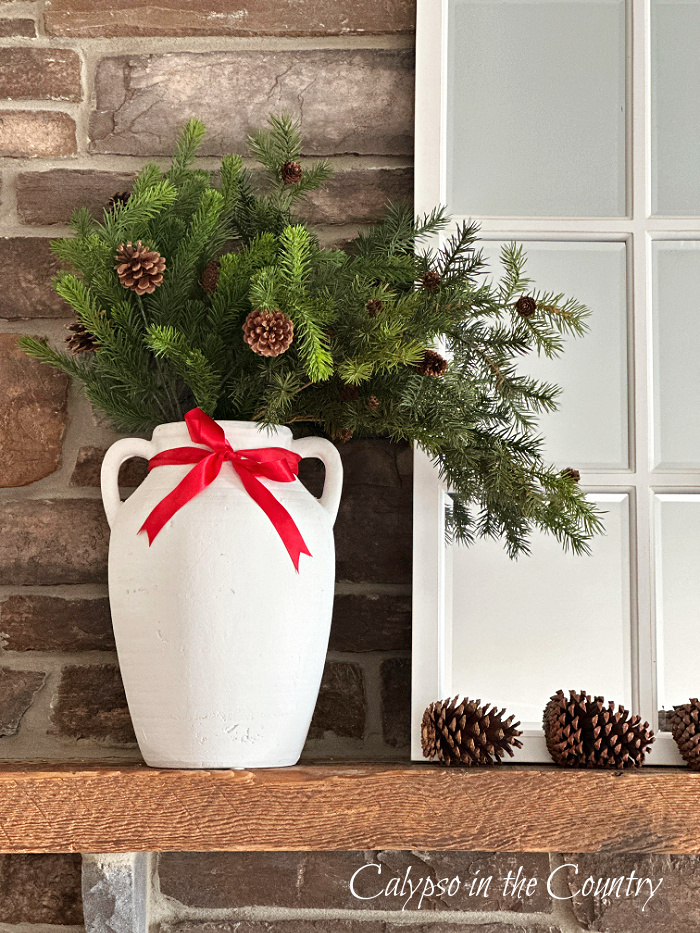 greenery in white vase with red bow next to mirror and pine cones on fireplace mantel