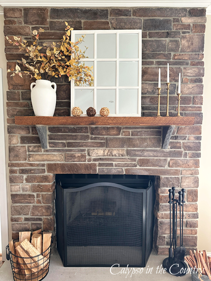 stone fireplace decorated for fall with golden foliage in white vase, white paned mirror, candles and wicker orbs