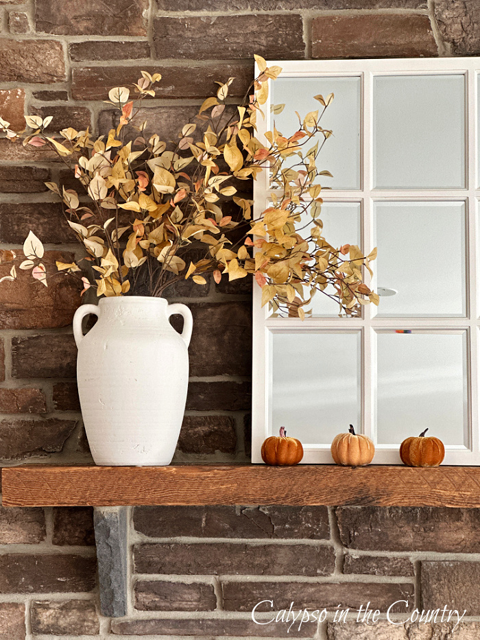 stone fireplace styled for fall with golden foliage in white vase, white paned mirror and velvet pumpkins - decorate a mantel for every season