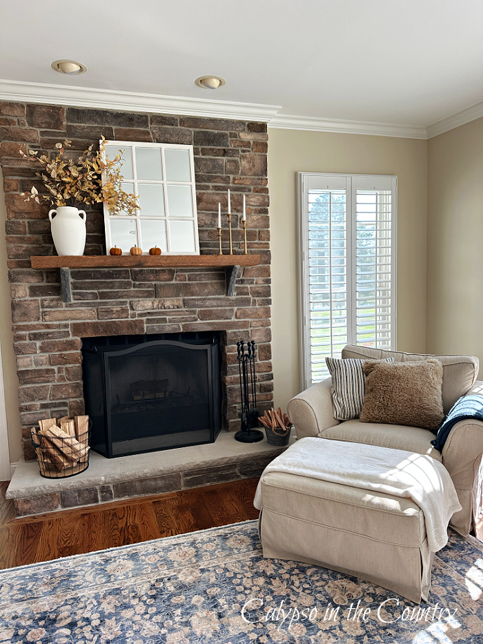 Family room with chair and ottoman and stone fireplace styled for fall with golden foliage in white vase, white paned mirror and velvet pumpkins - decorate a mantel for every season