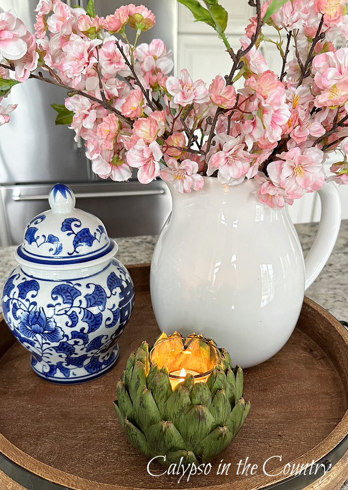 Artichoke candle holder on round tray with white pitcher of pink cherry blossoms and blue and white ginger jar