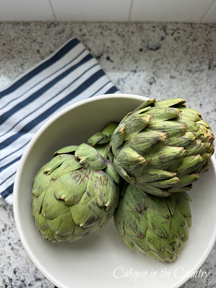 Three artichokes in white bowl next to blue and white striped towel