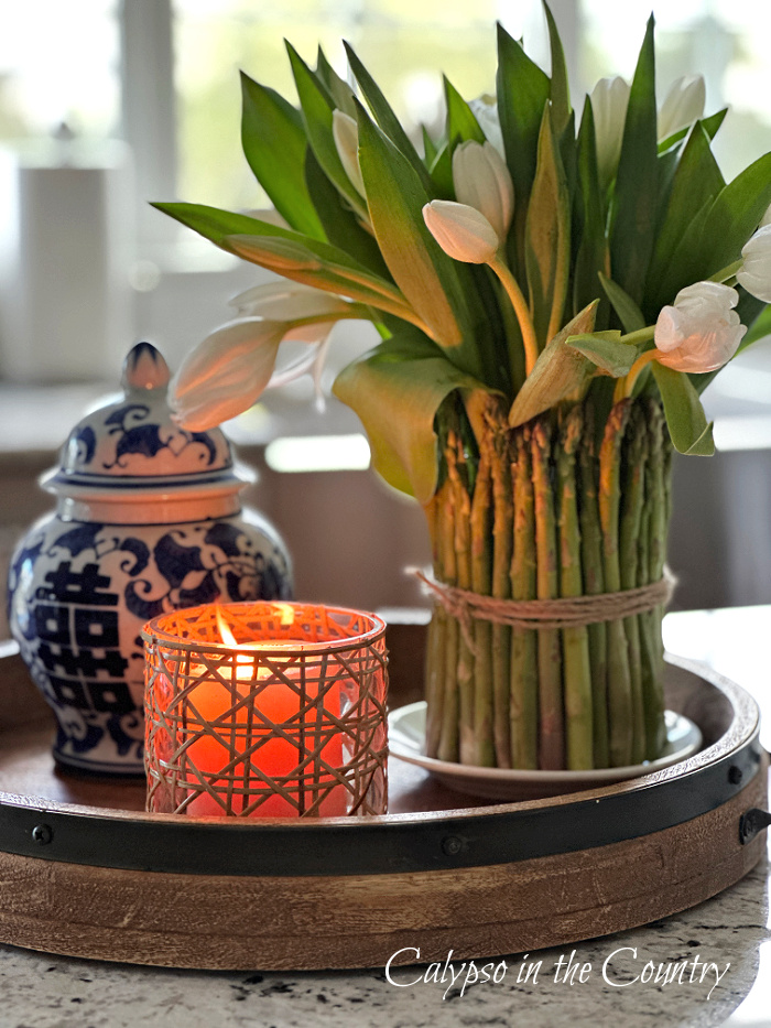 Asparagus vase with white tulips next to blue ginger jar and cane candle on round tray