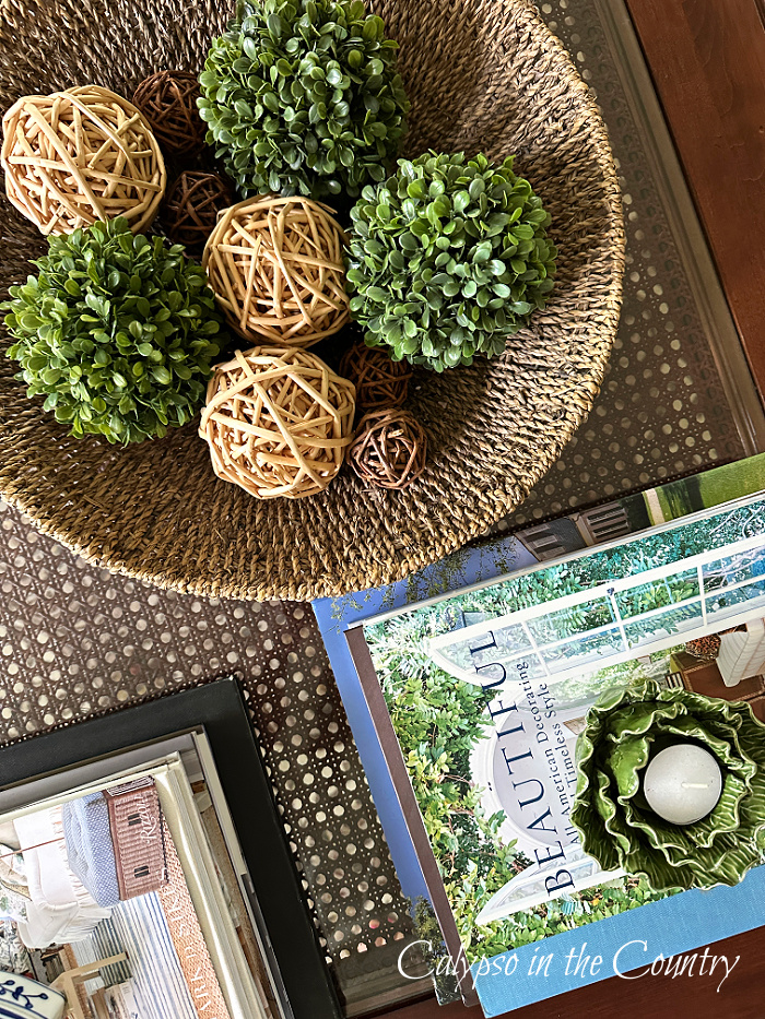 Woven bowl with green boxwood orbs and wicker orbs next to stacks of books on cane coffee table