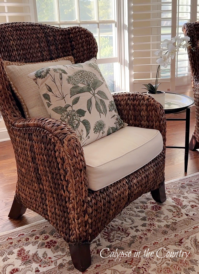 Green floral pillow on brown seagrass chair by window - Welcome May ideas