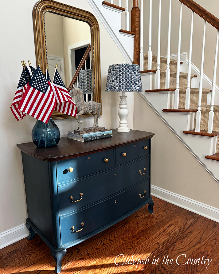 Flags in blue vase on blue dresser next to staircase in foyer - Decorating with American flags