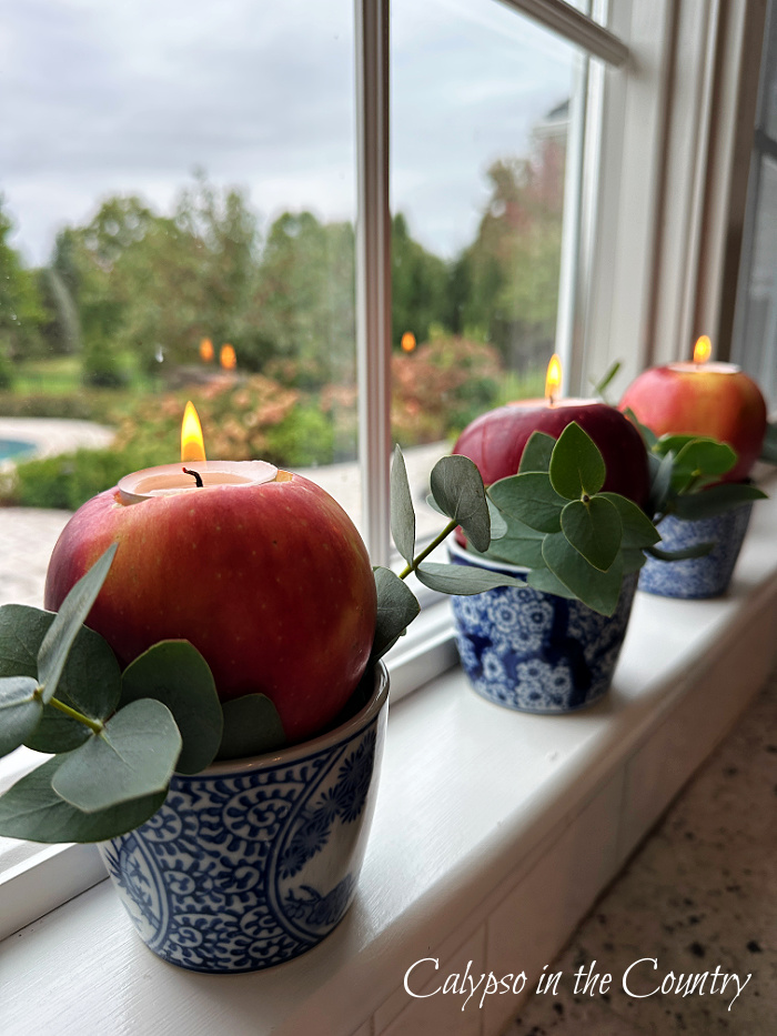 Three apples with candles in blue in white mini planters on window sill